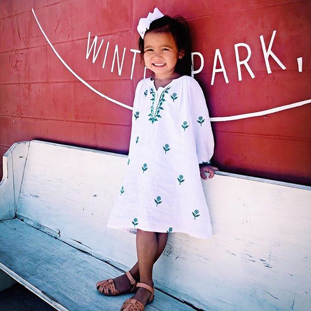 Little girl in her white Ava Dress standing against a concrete wall.