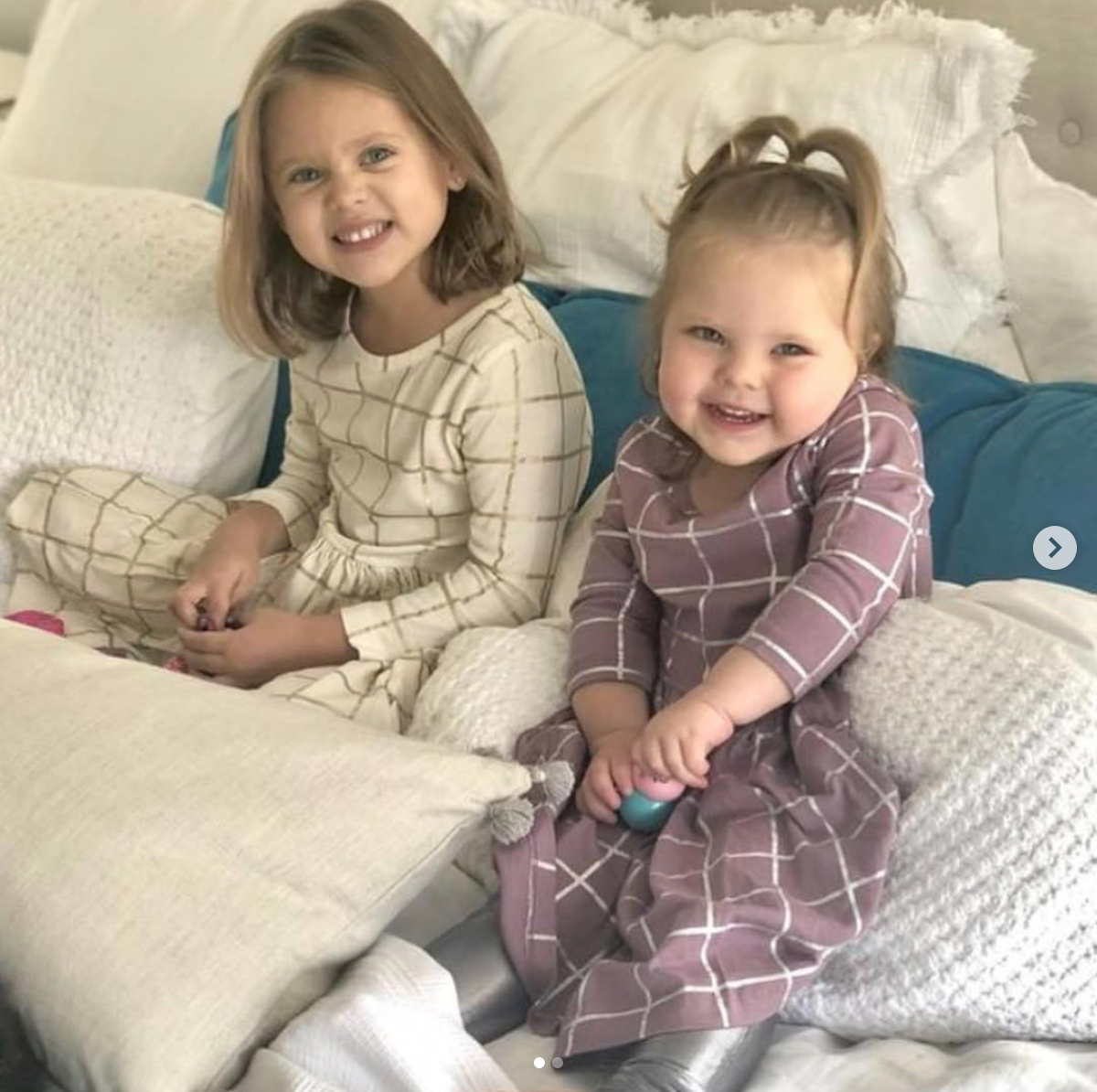 Little girls sitting in bed with their matching plaid long sleeve dresses - one in white and gold, the other in purple and silver.