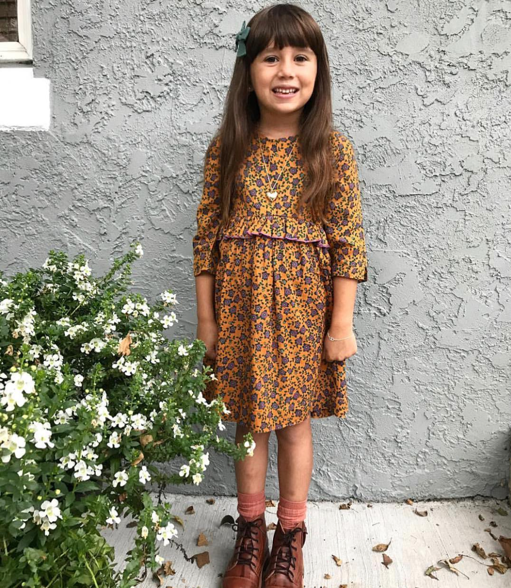 Young girl with brown hairs stands and smiles in her brown floral Pink Chicken Dress.