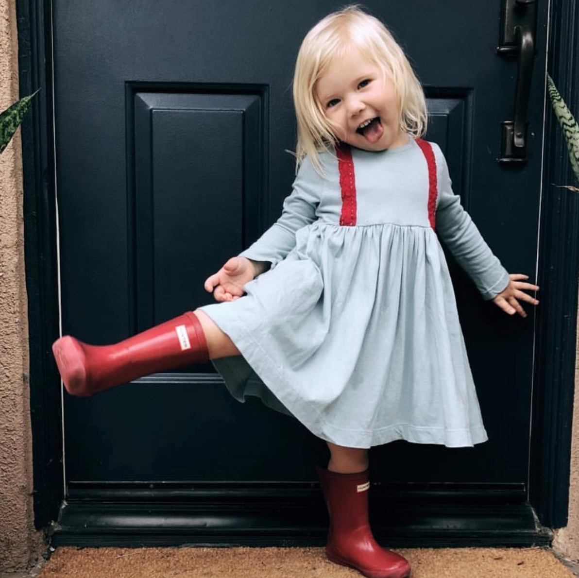 Little girl with blonde hair smiling big in her Pink Chicken gray and red dress. Wearing red rain boots too.