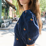 A woman wearing a navy blue eyelet puff-sleeve top with small embroidered floral details, photographed outdoors on a sunny city street.