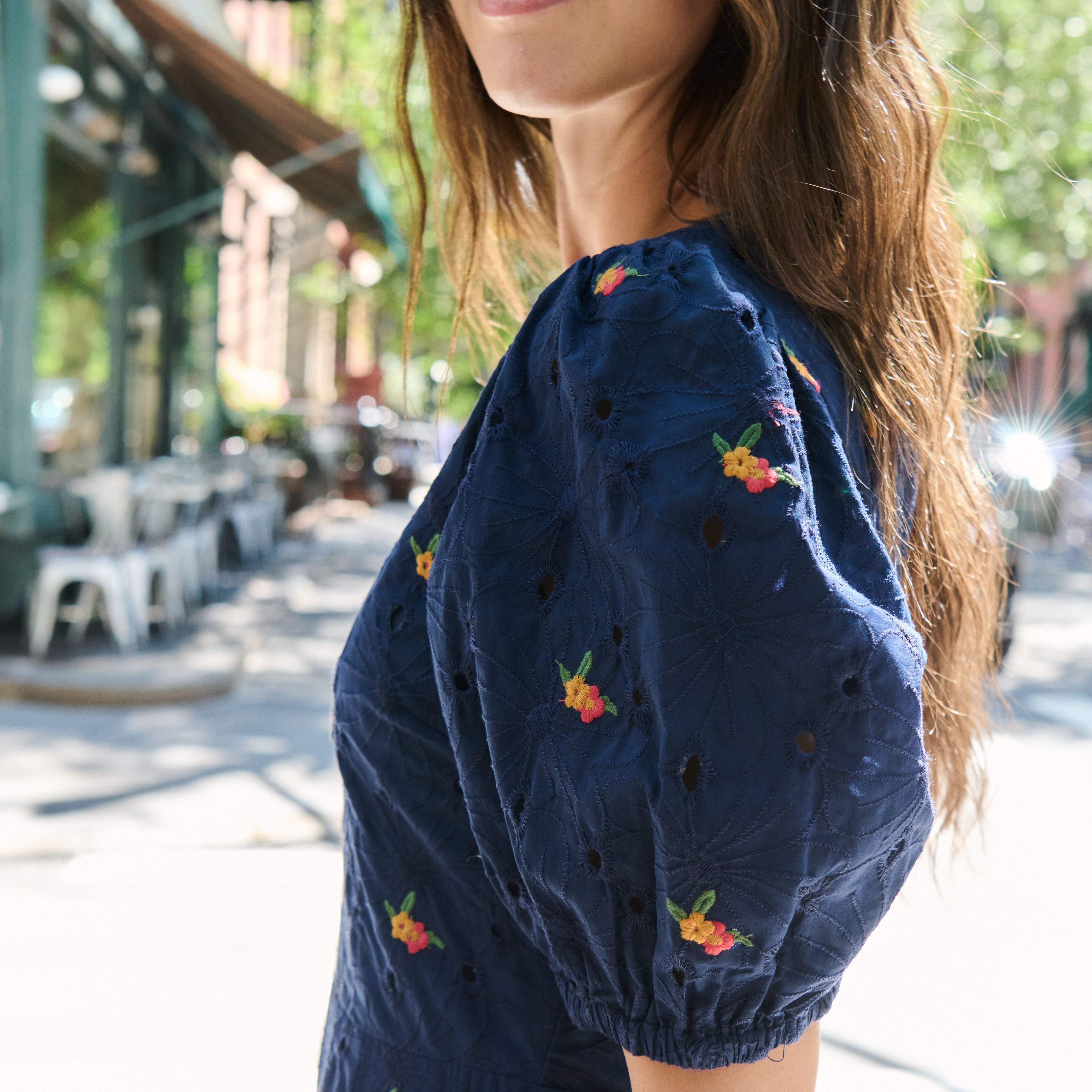 A woman wearing a navy blue eyelet puff-sleeve top with small embroidered floral details, photographed outdoors on a sunny city street.